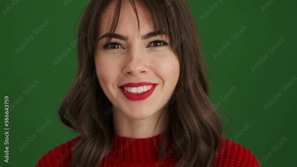 A close-up view of a cheerful woman in a red sweater is showing funny facial expression to the camera standing isolated over green background