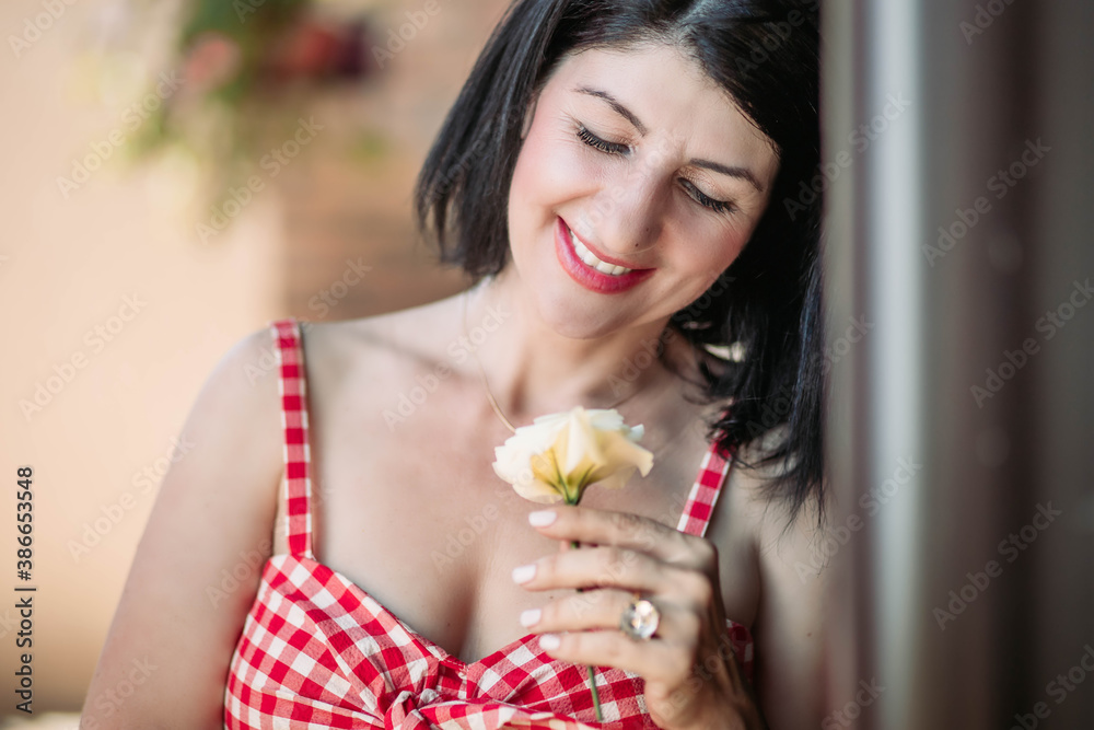 Happy brunette middle aged woman in red checkered dress holding white flower Happy brunette middle aged woman in red checkered dress holding white flower