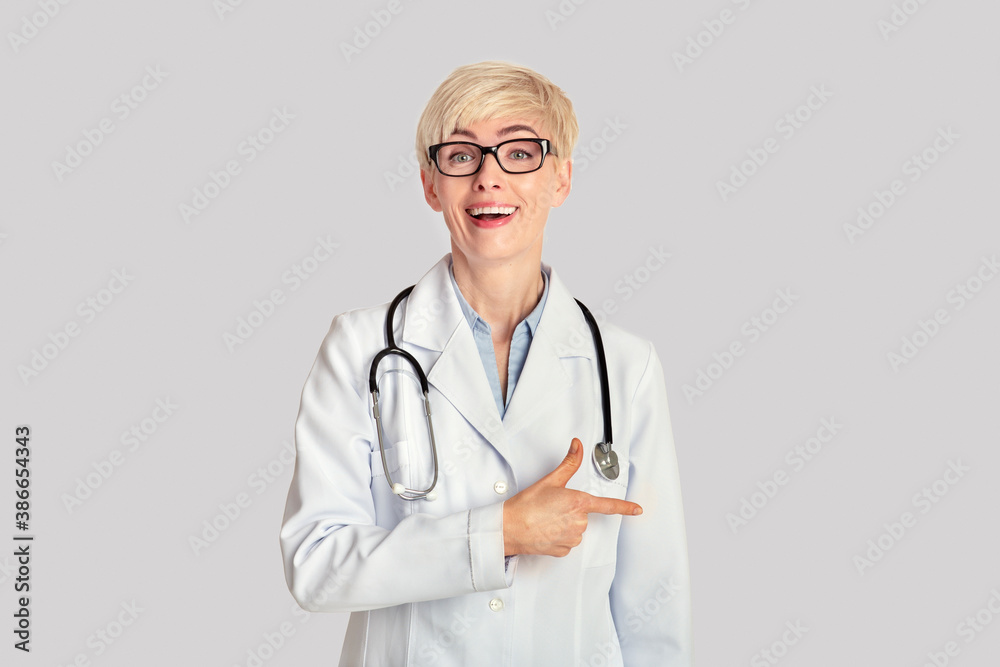 Shocked adult woman in white coat, glasses with stethoscope with open mouth points finger to empty space Shocked adult woman in white coat, glasses with stethoscope with open mouth points finger to empty space
