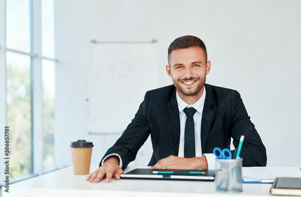 Stylish smiling businessman in elegant suit sitting at his desk in a bright modern office Stylish smiling businessman in elegant suit sitting at his desk in a bright modern office