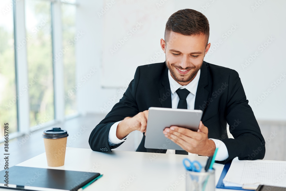 Happy successful businessman in elegant suite using tablet sitting in modern creative office Happy successful businessman in elegant suite using tablet sitting in modern creative office