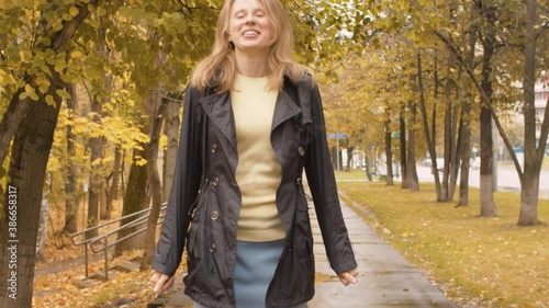 Happy woman jumping on the road near the park. Nice portrait of smiling lady. Yellow leaves on the trees. Autumn day in the city