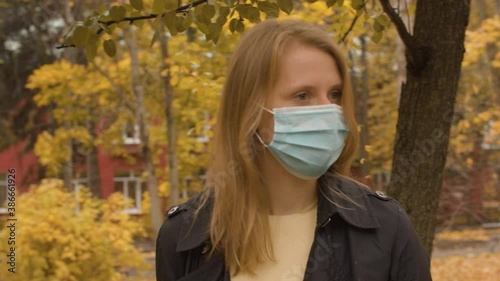 Portrait of young attractive woman in disposable protective mask standing in the autumn city. Yellow leaves on the trees, steadicam shot. Protection during viral epidemic.