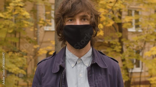 Close up portrait of young handsome man in black protective mask against autumn city. Yellow leaves on the trees. Autumn day, steadicam shot. Protection during viral epidemic.