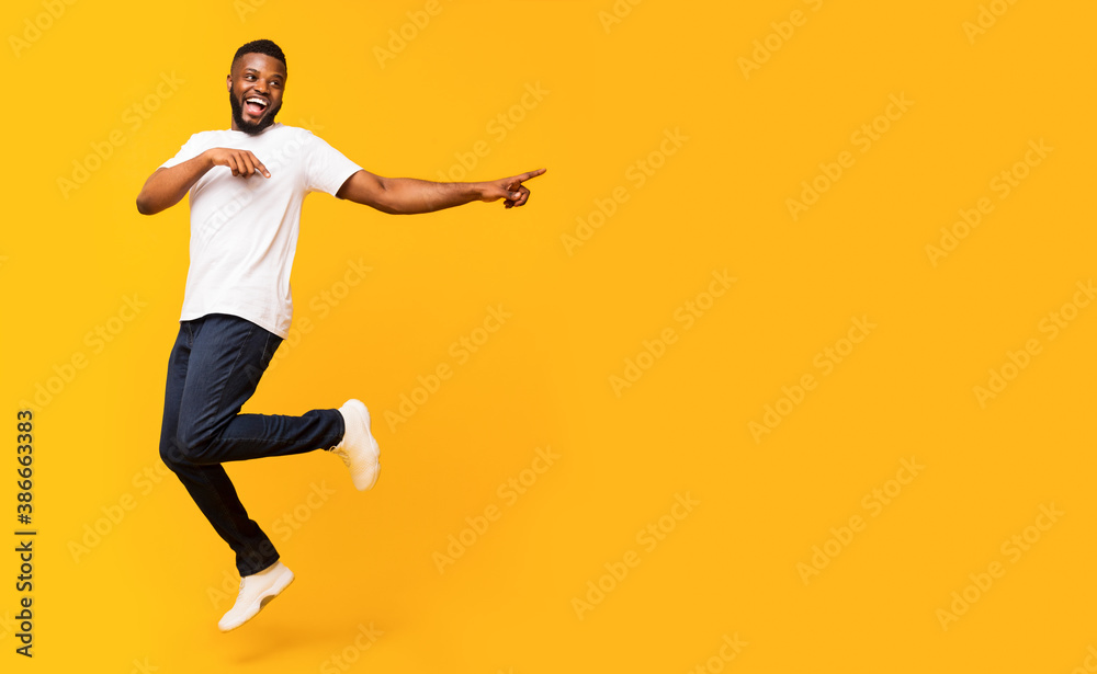 Joyful black guy jumping up and pointing aside - obrazy, fototapety, plakaty 