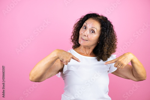Middle age woman wearing casual white shirt standing over isolated pink background looking confident with smile on face, pointing oneself with fingers proud and happy.