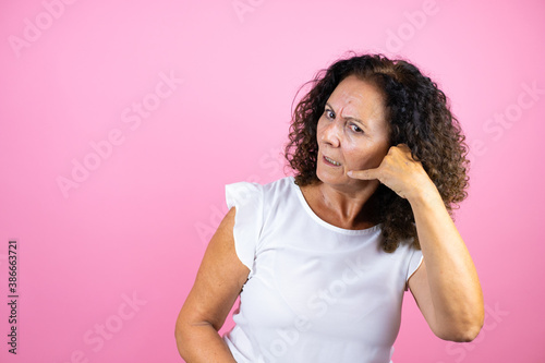 Middle age woman wearing casual white shirt standing over isolated pink background confused doing phone gesture with hand and fingers like talking on the telephone