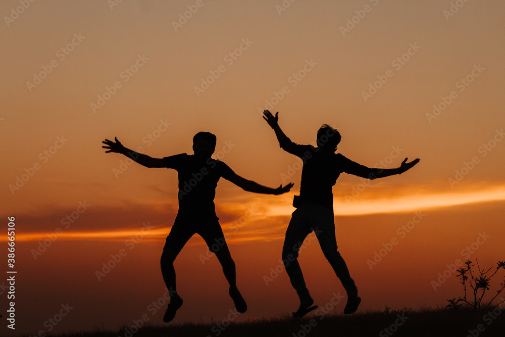 Fototapeta premium Silhouette of two Indian friends jumping with arms raised against the sky during the sunset 