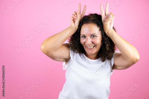 Middle age woman wearing casual white shirt standing over isolated pink background Posing funny and crazy with fingers on head as bunny ears, smiling cheerful