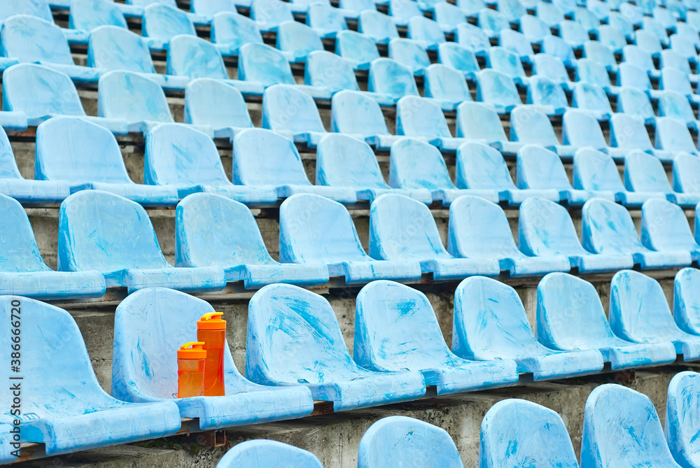 Naklejka premium Lots of empty seats in the stadium. Orange water bottle on the seat. Drink for athletes. Plastic seats are arranged in a row. Dirty and scratched blue fan seats.