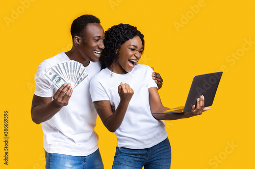 Emotional black couple celebrating win with laptop and cash