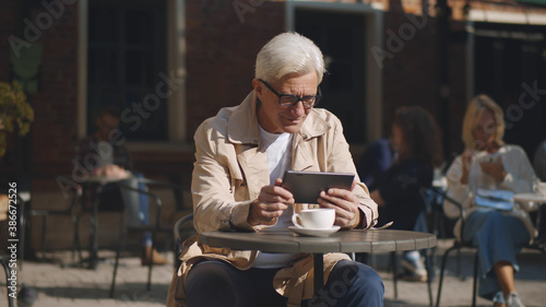 Smiling aged businessman using tablet and drinking coffee in outdoor cafe.