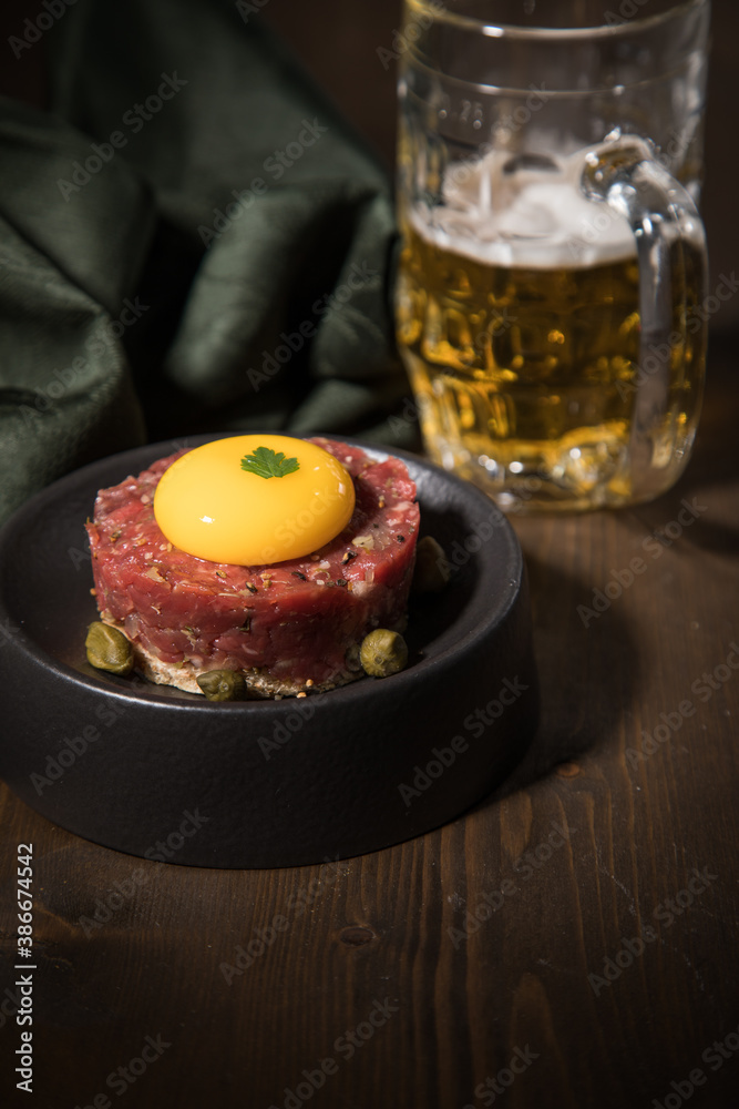Gourmet Rindertatar Beef Tatar roh aus Filet fettarm, Kapern, Eigelb, Zwiebeln auf Toast Brot und schwarz Teller mit Bier, grün Serviette und Holz Hintergrund dunkel Gourmet Rindertatar Beef Tatar roh aus Filet fettarm, Kapern, Eigelb, Zwiebeln auf Toast Brot und schwarz Teller mit Bier, grün Serviette und Holz Hintergrund dunkel