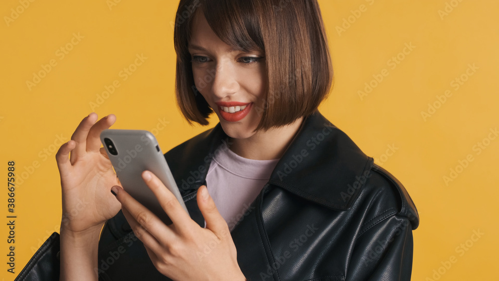 Naklejka premium Portrait of pretty girl with bob hair in casual wear happily surfes social media on smartphone isolated on yellow background