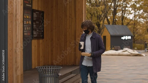 Young handsome man in a protective mask and gloves comes out of a fast food restaurant with a food box and a plastic cup in his hands. Autumn day in the city. Protection during viral epidemic.