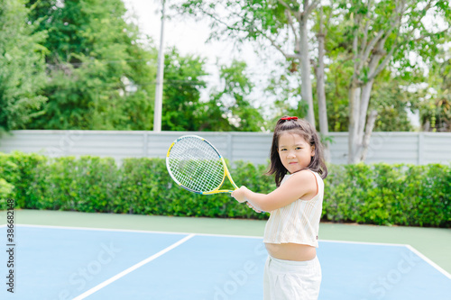 Little tennis player play tennis in court on summer.Confident asian child girl lose fist tooth with big smile she holding racquet for training ready to play tennis with trainer coach in sport club. © MIA Studio