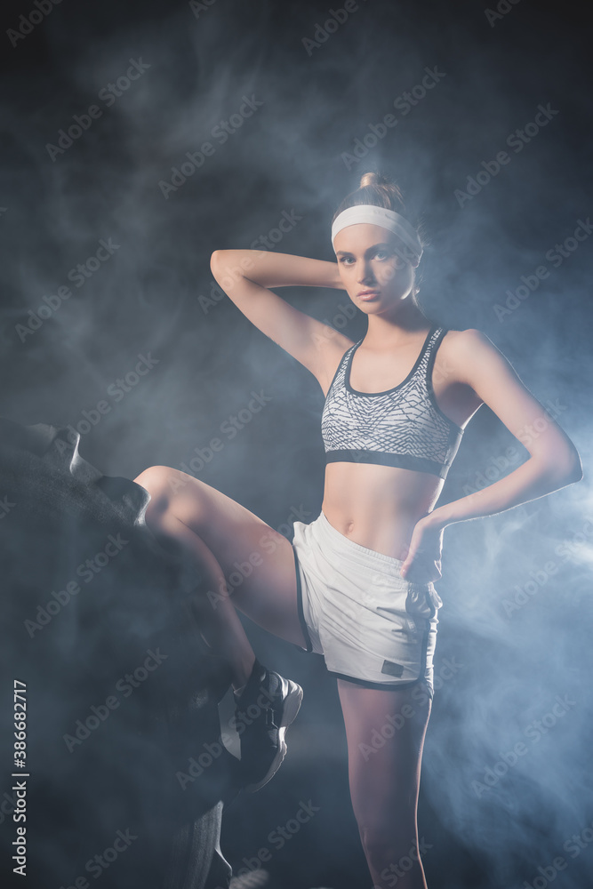 Obraz premium Selective focus of sportswoman looking at camera tear tire in sports center with smoke
