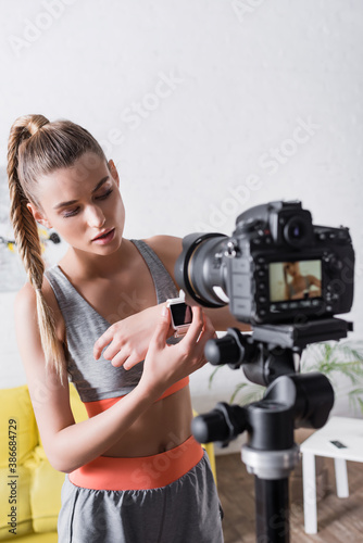 Selective focus of sportswoman showing smart watch at digital camera at home