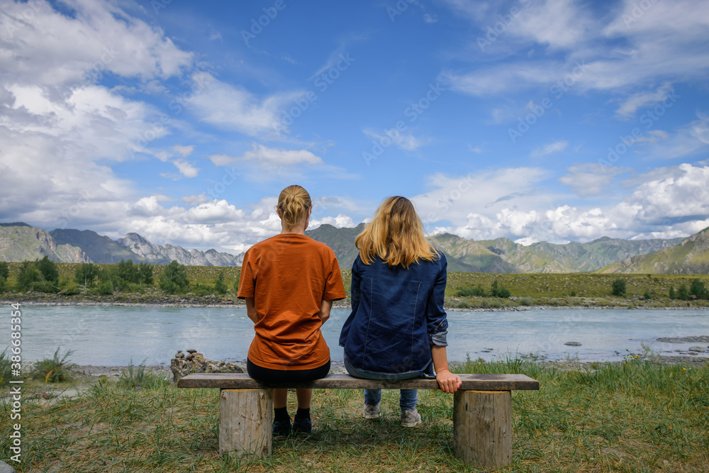 Two young women sitting on wooden bench, backs to the camera against beautiful landscape. Friends travel, admire magnificent view of mountain range under blue sky on sunny day. Two young women sitting on wooden bench, backs to the camera against beautiful landscape. Friends travel, admire magnificent view of mountain range under blue sky on sunny day.
