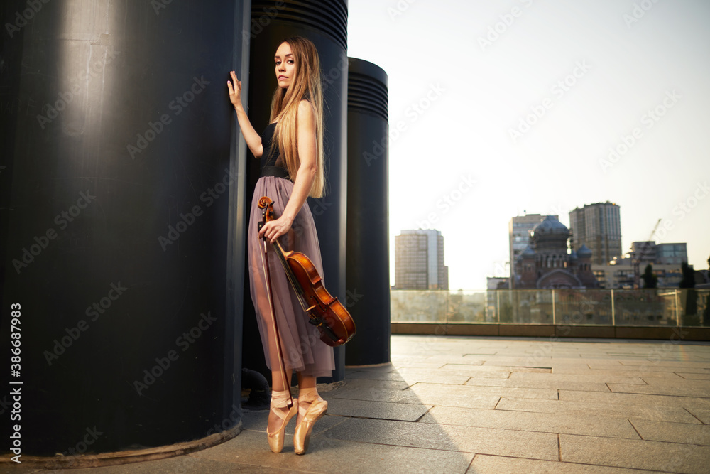 A beautiful adult woman with long hair posing with violin on pointe shoes outdoors in city. A beautiful adult woman with long hair posing with violin on pointe shoes outdoors in city.