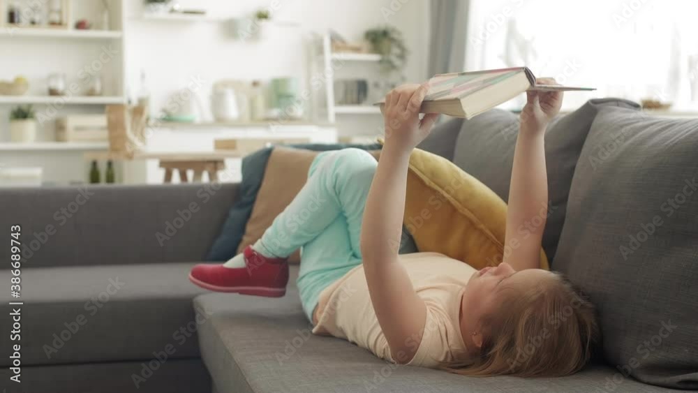 Lockdown of Caucasian girl with down syndrome lying on couch at home and reading interesting book Lockdown of Caucasian girl with down syndrome lying on couch at home and reading interesting book