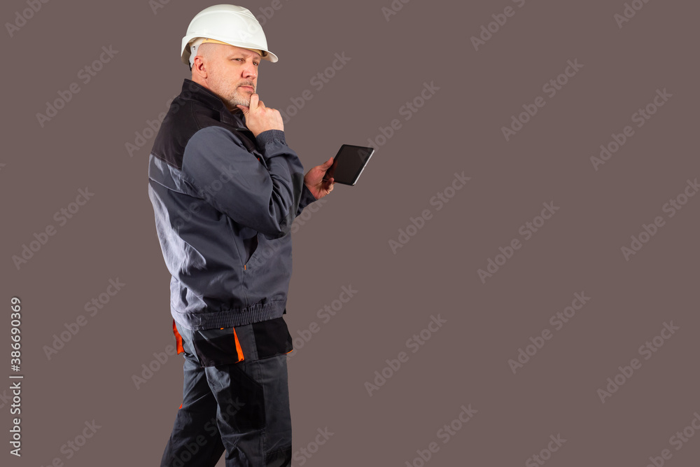 Thoughtful worker with a tablet in his hands. A man in a work uniform studies the details of the project. Repair and construction services. Portrait of the master on a brown background.