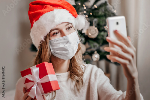 New Year's video calls. A young woman in a santa hat wearing a medical protective mask holds a red gift box and makes a video conference call. New Year video calls photo