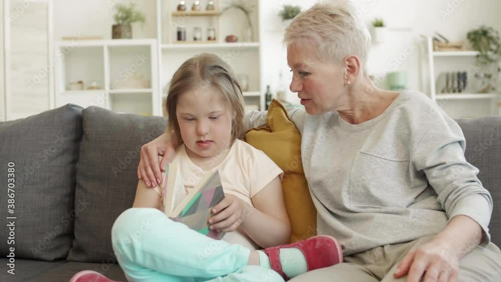 Lockdown of adorable Caucasian girl with down syndrome and her good-looking grandmother sitting together on sofa at home and reading book Lockdown of adorable Caucasian girl with down syndrome and her good-looking grandmother sitting together on sofa at home and reading book