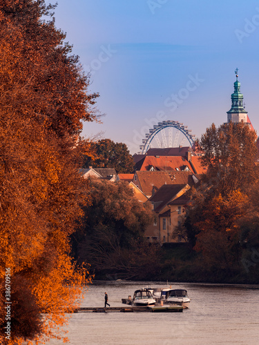 View over river with sports boats and colorful autumn trees with old town of Regensburg with ferris wheel in background n warm sunny october day