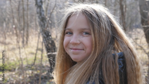 Portrait of happy small child against the blurred background of a forest in early spring. Smiling blonde girl looking into camera outdoor. Close up emotions of female kid with glad expression on face