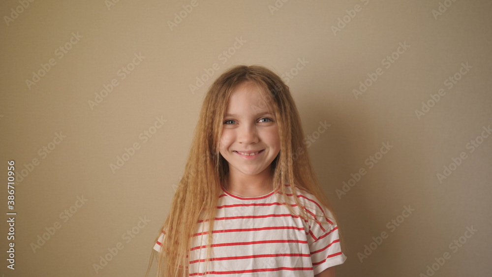 Little smiling girl with blonde hair looking into camera indoor. Close up emotions of beautiful kid with glad expression on her face. Portrait of happy female child against the background of wall Little smiling girl with blonde hair looking into camera indoor. Close up emotions of beautiful kid with glad expression on her face. Portrait of happy female child against the background of wall