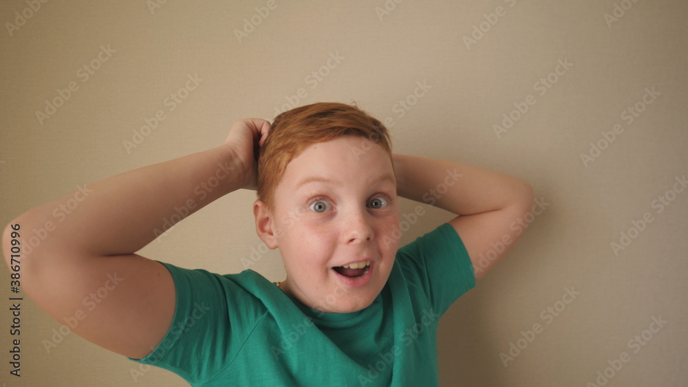Happy male ginger child with freckles looking into camera and raises hands rejoicing achievement inside. Portrait of little red-haired boy with positive emotions on his face indoor. Slow motion Happy male ginger child with freckles looking into camera and raises hands rejoicing achievement inside. Portrait of little red-haired boy with positive emotions on his face indoor. Slow motion