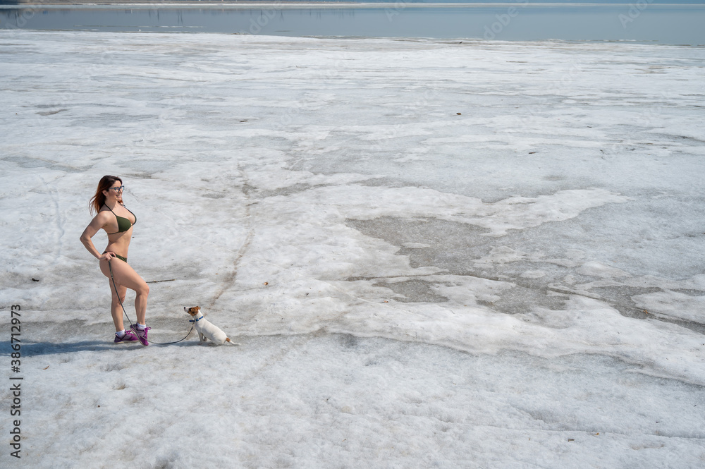 A red-haired woman in a bikini walks with a dog on a snowy beach