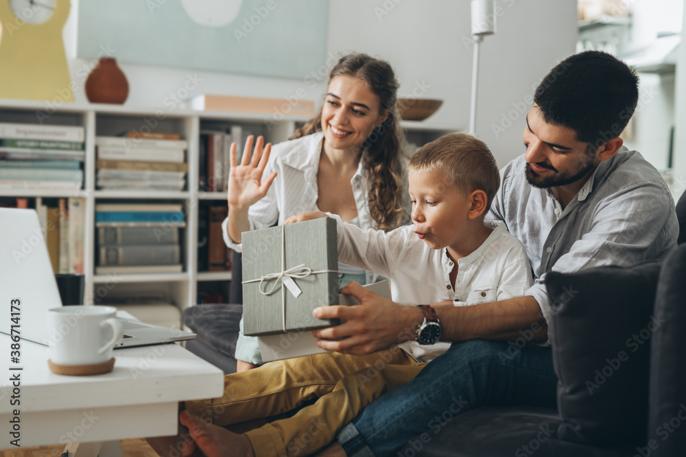 Obraz premium young happy family with one kid sitting on sofa and opening gifts while having video call on computer