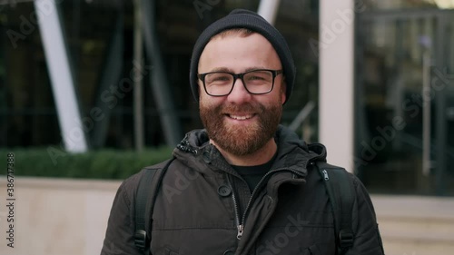 Close up view of joyful bearded guy in 30s posing while looking to camera. Portrait of handsome male person in glasseslaughing while standing at street. Concept of positive emotons. photo