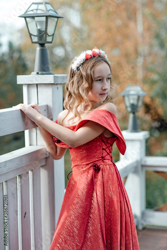 An attractive little girl in a long red dress walks along the open balcony. Yellowed autumn trees. Little princess. photo