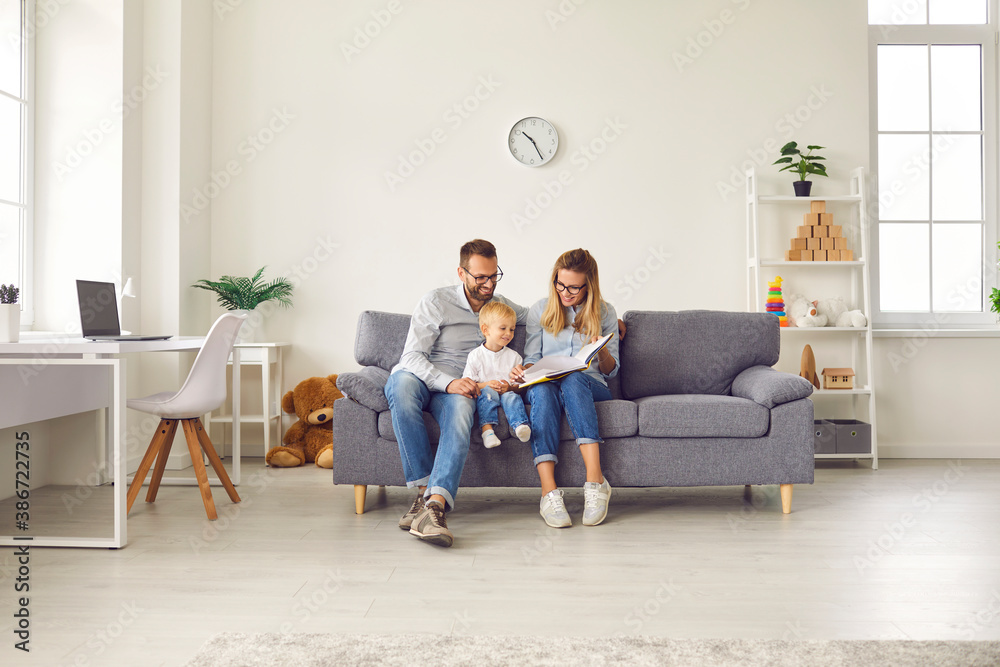 Happy young mom and dad sitting on comfy couch at home reading book together with their little son Happy young mom and dad sitting on comfy couch at home reading book together with their little son