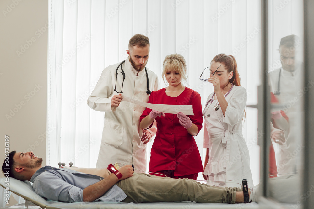Training with interns. A young blonde doctor gives a lecture to interns in the office, discussing the patient's cardiogram. The patient lies on the side of the couch. Medicine, education Training with interns. A young blonde doctor gives a lecture to interns in the office, discussing the patient's cardiogram. The patient lies on the side of the couch. Medicine, education
