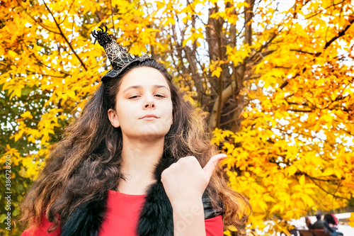 Girl brunette in a fairytale image in an autumn park. A girl in a red dress and a black vest against a background of yellow leaves points with a finger to the empty space. copy space