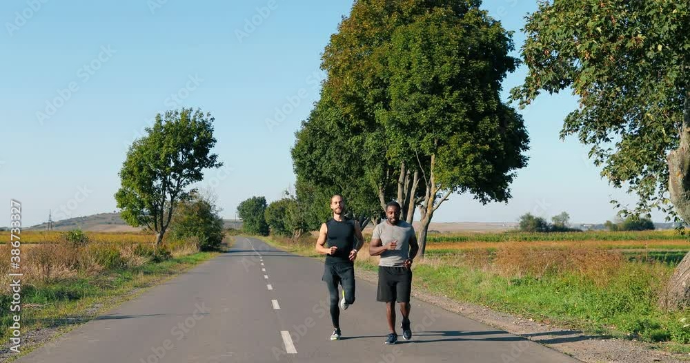 African American and Caucasian young sporty men friends joggers running on road early in the morning in countryside. Males runners. Mixed-races sportswomen jogging in nature. African American and Caucasian young sporty men friends joggers running on road early in the morning in countryside. Males runners. Mixed-races sportswomen jogging in nature.