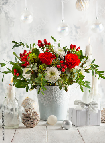 Festive winter flower arrangement with red roses, white chrysanthemum and berries in vase on table decorated for holiday. Christmas or New Year concept.