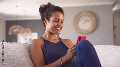 Cheerful young african woman using smartphone while sitting on couch. Black smiling woman using app on cellphone at home. Beautiful girl relaxing while chatting on mobile phone and looking at camera. 