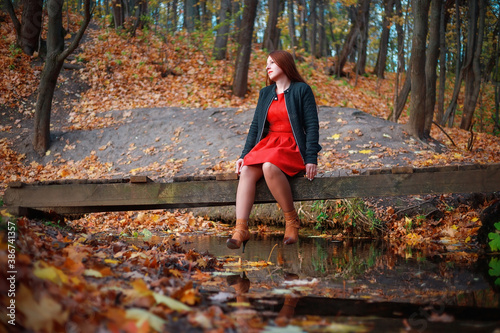 The girl in the red dress. Autumn park. The model is sitting on a wooden bridge. Wooden bridge over the stream.
