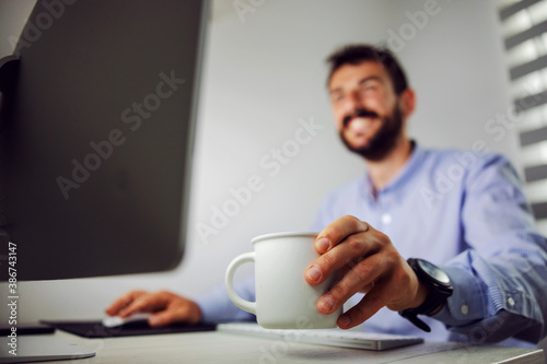 Closeup of smiling businessman using computer and holding mug with coffee while sitting in his office. Selective focus on mug.