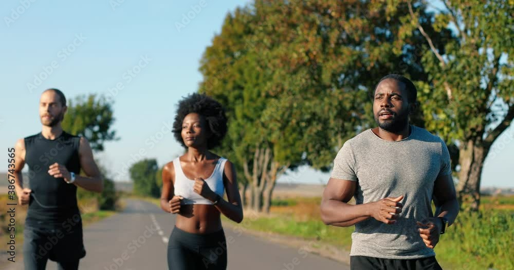 Mixed-races young males and female joggers running on countryside road in summer. African American slim sporty woman and multi ethnic men jogging outdoor. Runners workout together. Mixed-races young males and female joggers running on countryside road in summer. African American slim sporty woman and multi ethnic men jogging outdoor. Runners workout together.
