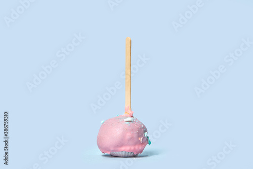 Chocolate dessert in pink glaze with colored sprinkles, dragees. Round pastry on a wooden stick. Pink icing. Blue background.
