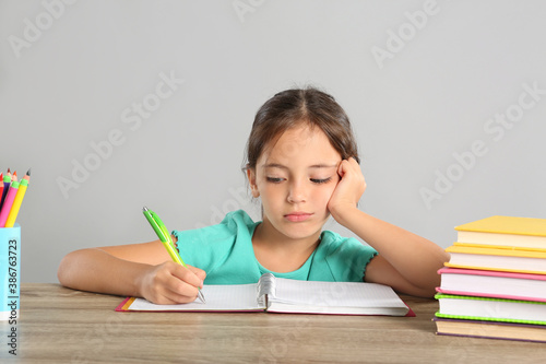 Bored little girl doing homework at table on grey background