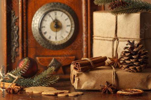 Gifts close-up in craft paper with winter spices and fir branches. Vintage clock on the mantelpiece showing midnight in the background.