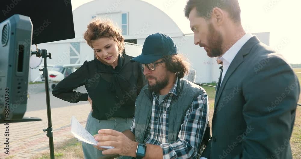 Portrait of Caucasian member of film crew talking outdoors on set. Male and female chatting with movie director on backstage. guy in cap discussing script with actors. Filmmaking. Shooting concept