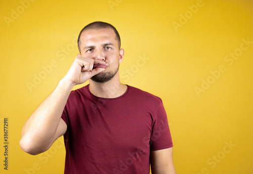 Russian man wearing basic red t-shirt over yellow insolated background mouth and lips shut as zip with fingers. Secret and silent, taboo talking